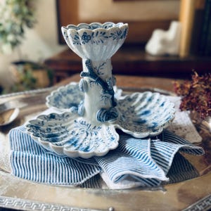May include: A white porcelain serving dish with a central pedestal and three shell-shaped compartments. The piece features intricate blue floral patterns and raised blue branch and shell motifs on the central column and scalloped edges.