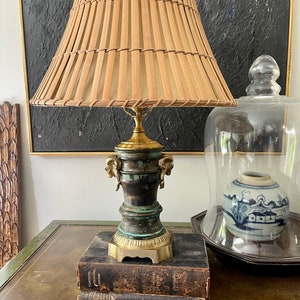 May include: A brass table lamp with a green patina finish and a woven bamboo shade. The lamp is sitting on a stack of three books.