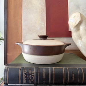 May include: A vintage, two-toned brown and cream-colored casserole dish with a lid. The dish is sitting on top of two antique books with the title "History of English Plate" visible. The background features a neutral color palette.