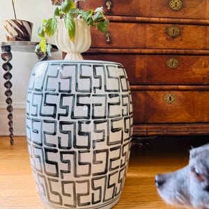 May include: A white ceramic garden stool with a repeating black geometric pattern. The stool is cylindrical and sits on a wooden floor. A plant in a white vase sits on top of the stool.