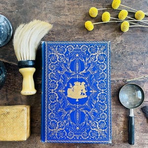 Puede incluir: Un libro antiguo con una cubierta azul y un diseño floral dorado, una brocha de afeitar, una pequeña caja dorada, una lupa y flores amarillas sobre una superficie de madera. El libro tiene un emblema dorado en el centro.