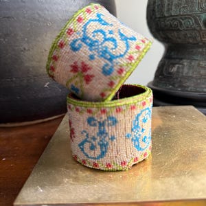 May include: Two beaded napkin rings. The rings are beige with blue floral designs and red accents. The edges are trimmed with green beads. One ring is stacked on top of the other.