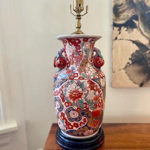 May include: A white porcelain table lamp with a floral design in red, blue, gold, and green. The lamp has two handles shaped like apples and a black base.