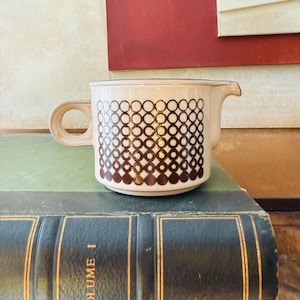 May include: A vintage cream-colored creamer with a brown geometric pattern. The creamer has a handle and a spout. It is sitting on a green book with the words "VOLUME I" in gold lettering.
