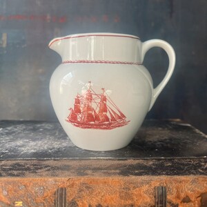 May include: A white ceramic creamer with a red ship design. The creamer has a handle and a spout. It is sitting on top of a brown leather-bound book.
