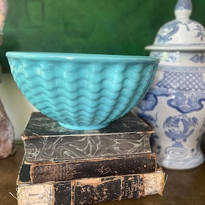 May include: A turquoise blue ceramic bowl with a wavy textured design sits on top of a stack of three antique books. The bowl is a decorative piece and is not being used for serving.
