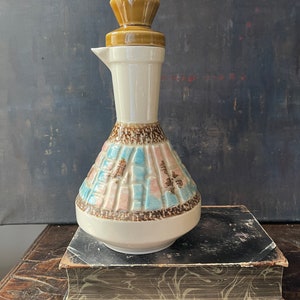 May include: A vintage ceramic pitcher with a tan lid and a white body. The lower portion features a geometric pattern in blue, pink, and brown. The pitcher is displayed on a stack of old books, showcasing a retro design.