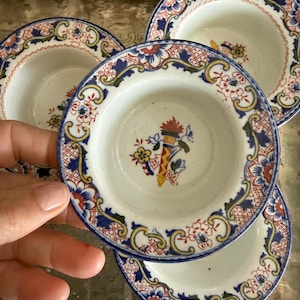 May include: A set of four small, white ceramic bowls with a detailed floral and geometric pattern in blue, red, and yellow. The bowls have a blue rim and a central floral design. The bowls are likely for serving small portions.