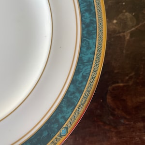 May include: White ceramic plate with a teal and gold border. The border features a marbled teal band, framed by gold lines and a repeating geometric pattern. A small blue floral design is visible.
