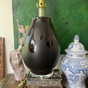 May include: Black ceramic table lamp with a teardrop-shaped base and a gold-tone finial. The lamp is sitting on a stack of antique books.