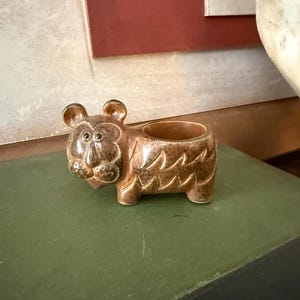 Puede incluir: Un macetero de cerámica marrón con forma de tigre. El macetero tiene una cara detallada, orejas y un patrón de pelaje texturizado. La abertura es un cuenco pequeño y redondo. El macetero está sobre una superficie verde.