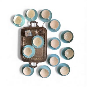 May include: An overhead shot of a vintage silver serving tray with handles, surrounded by light blue saucers and cream-colored teacups. The arrangement includes a small decorative box and dried sprigs.