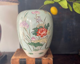 May include: A light blue ceramic vase with a floral design featuring pink and red lotus flowers, green leaves, and two small birds. The vase is sitting on top of two old books.