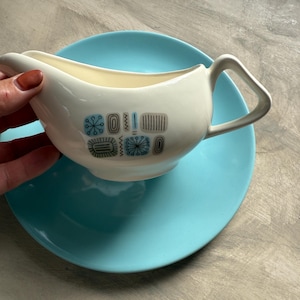 May include: A white ceramic gravy boat with a blue and black geometric pattern, resting on a turquoise plate. The gravy boat has a handle and a spout. The plate is round and has a smooth surface. The background is a neutral gray.