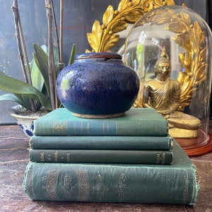 May include: A dark blue ceramic jar with a brown rim sits atop a stack of four antique green books. The books have gold lettering and are arranged on a wooden surface. A golden Buddha statue is visible in the background.