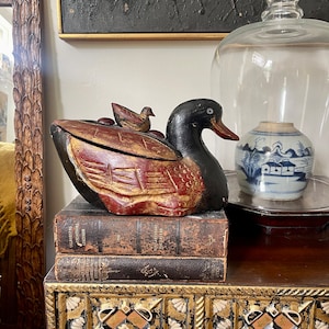 May include: A carved wooden duck-shaped box with a black head, red and gold body, and a small duckling on top. The box sits atop two antique books on a gold and black decorated chest.