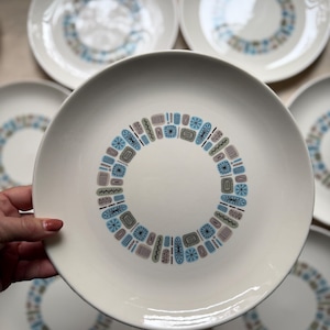 May include: White ceramic plates with a mid-century modern design. Each plate features a central ring of geometric shapes in blue, brown, and gray. The plates are round and have a simple, clean aesthetic, suitable for serving meals.
