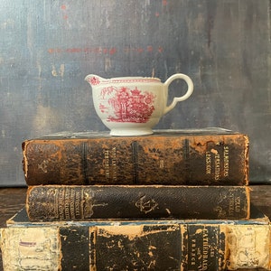 May include: A stack of three antique books with leather covers. The top book has the title "SALMONSENS K CONVERSATIONS LEKSIKON" in gold lettering. The middle book has the title "NORDLANDSBAAN PERLEDE ERNE" in gold lettering. The bottom book has the title "LES CATHEDRALES DE FRANCE" in gold lettering. A small white creamer with a pink floral design sits on top of the books.