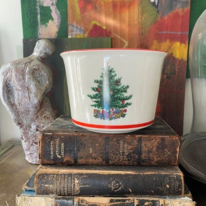 May include: A white ceramic planter with a red rim features a Christmas tree design with a red and green color scheme. The planter is sitting on top of a stack of three vintage books with brown leather covers.