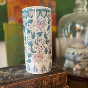 May include: A white ceramic vase with a floral pattern in shades of green, blue, and pink. The vase has a cylindrical shape and a slightly flared rim. It is sitting on top of a stack of old books.