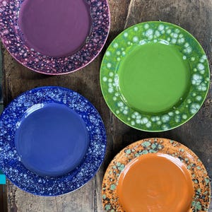 May include: Four ceramic plates with speckled glaze in different colors: purple, green, blue, and orange. The plates are arranged on a wooden surface.