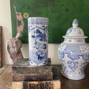 Puede incluir: Un jarrón cilíndrico de porcelana azul y blanca con un diseño de grullas, sobre una pila de libros antiguos. A la derecha hay un tarro con tapa a juego. El jarrón mide unos 30 cm de alto.