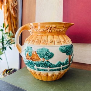 May include: A vintage ceramic pitcher with a landscape design. The pitcher features a cream-colored body with a detailed scene of trees, a house, and a white fence. The top and bottom are a warm orange color with floral and bow details.