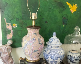 May include: A pink and white porcelain table lamp with a bird and floral design. The lamp is on a stack of books and next to a blue and white porcelain ginger jar.