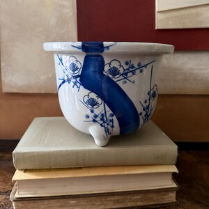 May include: A white ceramic planter with a wide rim and three small feet. The exterior features a hand-painted blue design of thick flowing brushstrokes and delicate plum blossom motifs. The vessel has a glossy finish and a rounded, tapered shape.