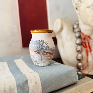 May include: Small, off-white ceramic vase with a brown rim and a painted landscape scene featuring a tree. The vase is cylindrical and sits on a stack of books. The background includes a red and white striped surface and a statue.