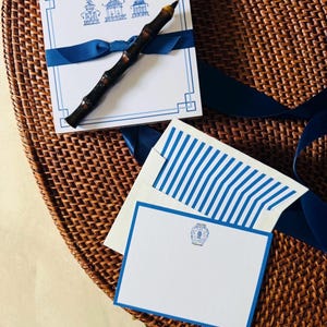 May include: Stationery set featuring white notepads and cards with blue accents. The notepads have a blue border and a pagoda design. A dark bamboo pen rests on the notepad, secured with a blue ribbon. The cards have blue striped envelopes.