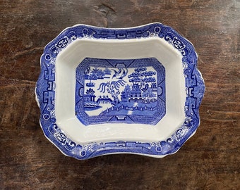 May include: A rectangular, white ceramic dish with a blue Willow pattern design. The dish has a scalloped edge with a blue border. The center features a detailed landscape scene with buildings, trees, and a bridge.