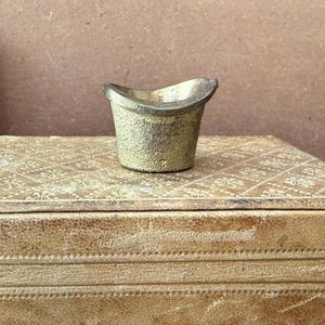 May include: A small, gold-colored, top hat-shaped container with a textured surface. The container sits atop a box with a patterned design. The box and container are in a warm, neutral color palette.