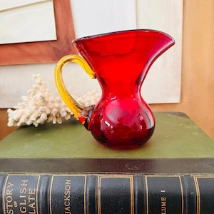 Może przedstawiać: Mały, zabytkowy, czerwony szklany dzbanek z żółtym uchwytem. Dzbanek ma zaokrągloną podstawę i rozszerzoną krawędź. Uchwyt jest zakrzywiony i przymocowany do boku dzbanka. Dzbanek stoi na stosie książek.