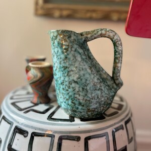 Puede incluir: Una jarra de cerámica turquesa con un esmalte moteado y un asa marrón. La jarra está sentada sobre un pedestal blanco con un patrón geométrico negro.