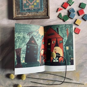 Puede incluir: Un libro abierto con ilustraciones coloridas de casas y figuras, junto a un tablero de juego enmarcado en madera con un diseño geométrico. Bloques de madera dispersos en varios colores y tallos de flores secas completan la escena.