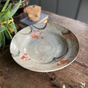 May include: A ceramic plate with a white glaze and a hand-painted design of orange and brown abstract shapes. The plate has a slightly raised rim and a speckled surface.