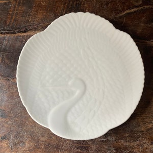 May include: A white ceramic plate with a swan design. The plate has a scalloped edge and a raised relief pattern of feathers and the swan's neck. The plate is round and sits on a dark wooden surface.