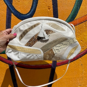 May include: White leather shoulder bag with a patchwork design featuring reptile skin patterns in shades of brown, beige, and cream. The bag has a tassel and a thin white strap. The bag is held against a colorful graffiti background.