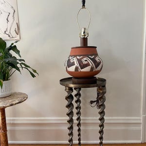 May include: A ceramic lamp with a geometric pattern sits on a table with three twisted legs. The lamp has a brown base and a white and brown geometric pattern. The table is made of metal and has a brown finish.