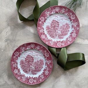 May include: Two red and white decorative plates with a landscape design. The plates have a scalloped edge and a floral border. A green ribbon is draped around the plates.