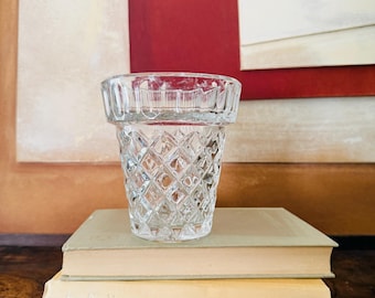 Puede incluir: Un vaso de cristal transparente con un patrón de diamantes en la parte inferior y un borde liso con nervaduras verticales. El recipiente tiene una abertura ensanchada y una base gruesa, mostrando una superficie transparente y reflectante con detalles geométricos.