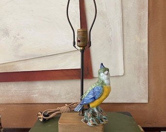 May include: A vintage table lamp featuring a colorful ceramic parrot perched on a wooden base. The lamp has a dark metal frame and a brown cord. The lamp sits on a stack of books with the title "History of English Plate".