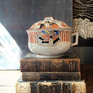 May include: A white ceramic lidded bowl with a handle, featuring a Japanese warrior design in orange, blue, and yellow. The bowl is set atop a stack of antique books with worn, leather-bound covers. The background is a dark, textured surface.