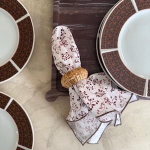 May include: Overhead shot of a dining table setting. White plates with brown geometric borders are arranged with a matching napkin and woven napkin ring. A dark brown tray and a floral patterned napkin complete the setting.