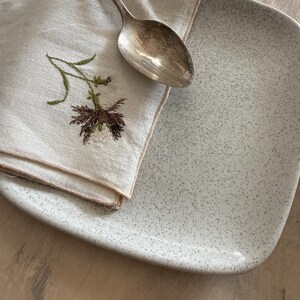 May include: A light gray speckled ceramic plate with rounded corners. A folded linen napkin with embroidered floral detail and a silver spoon rest on the plate. The napkin features a brown and green floral design.