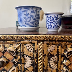 Puede incluir: Dos pequeñas tazas de porcelana azul y blanca con motivos florales. La taza más grande se asienta sobre un platillo pequeño. Las tazas están sobre una superficie de madera oscura con detalles dorados y negros ornamentados.