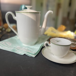 Puede incluir: Un juego de té de porcelana blanca con ribete verde, que incluye una tetera, una taza y un platillo. La tetera tiene un diseño acanalado y un pico curvo. La taza y el platillo también son blancos con ribete verde y un diseño floral.