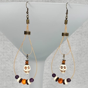 May include: A pair of dangle earrings featuring a skull design. The earrings have a gold-colored wire frame with a small skull pendant and beads in white, purple, and brown. The earrings are designed for pierced ears.