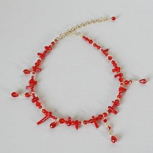 May include: A red beaded necklace with a gold-colored chain and accents. The necklace features various sizes and shapes of red beads, including small round beads and larger, irregular-shaped beads. Some beads dangle from the necklace.